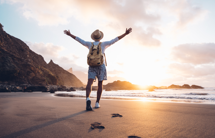 Man wearing a backpack and walking along beach with arms outstretched, wearing touristy hat